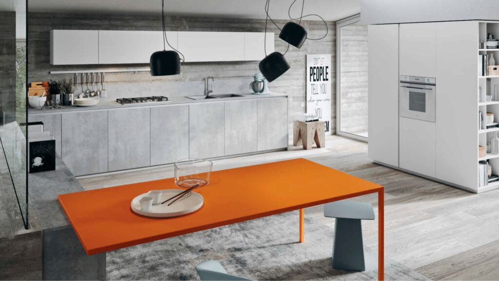Cocina moderna con mobiliario blanco y gris cemento, mesa naranja vibrante y diseño abierto con luz natural.